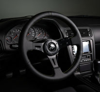 A black leather racing steering wheel with green contrast stitching and a black center hub, displayed on a black stand against a white studio background.