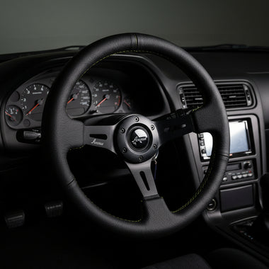 A black leather racing steering wheel with green contrast stitching and a black center hub, displayed on a black stand against a white studio background.