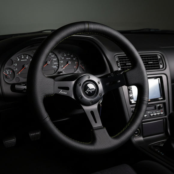 A black leather racing steering wheel with green contrast stitching and a black center hub, displayed on a black stand against a white studio background.